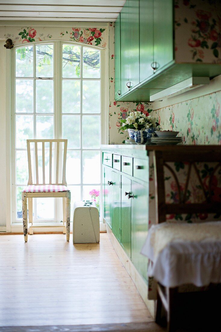 Weisser Stuhl vor raumhohen Sprossenfenstern, seitlich Einbauschränke mit grünen Fronten in ländlicher Küche