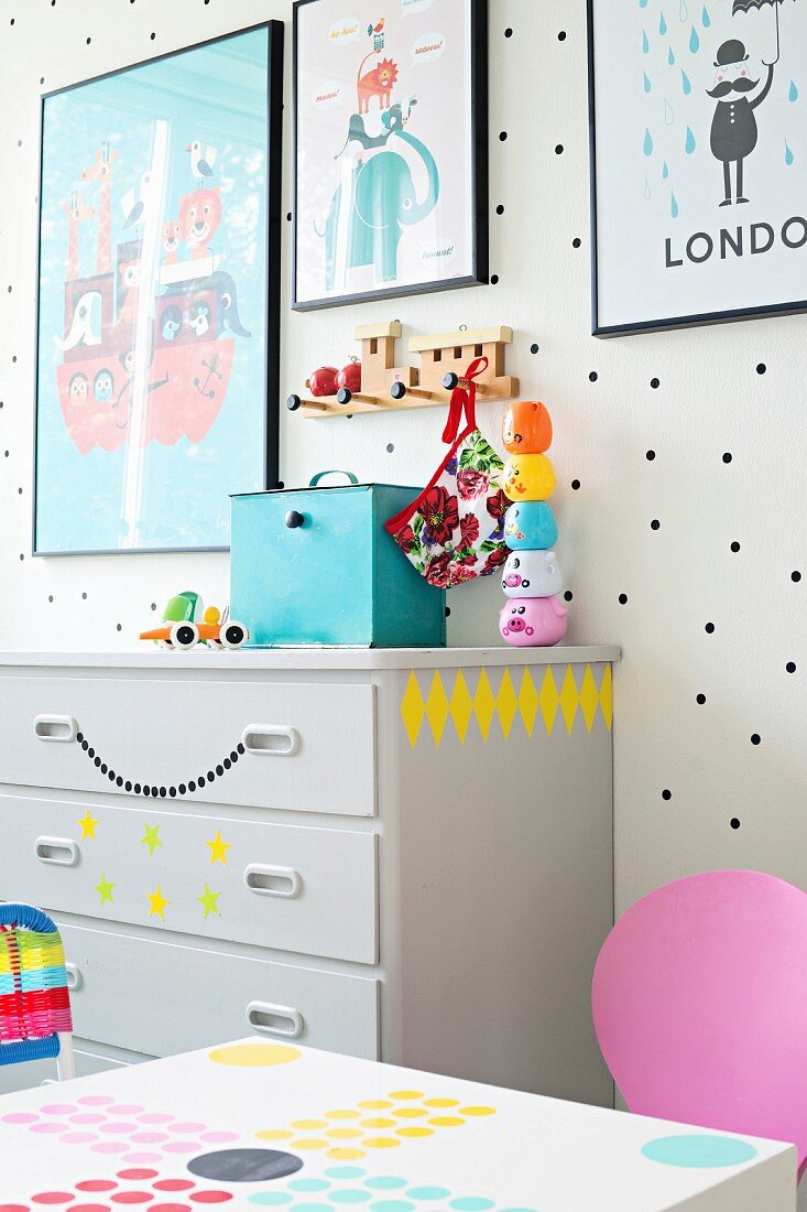 Framed posters on white wall with black polka dots and toys on grey chest of drawers with decorative stickers