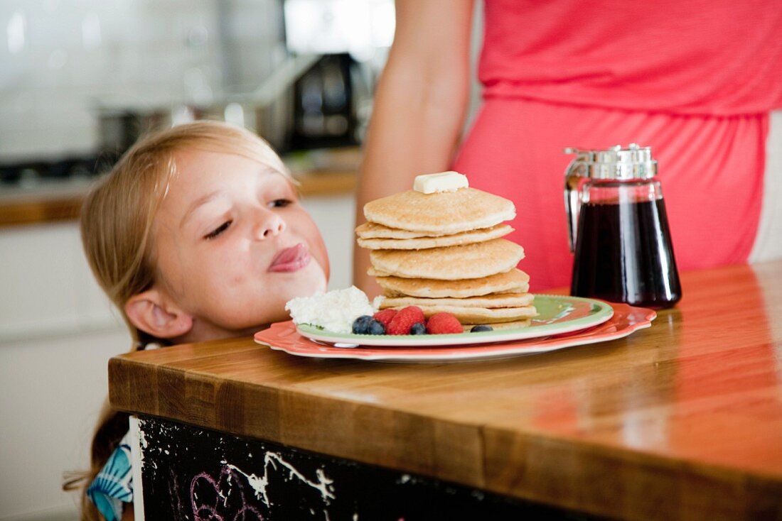 A mischievous little girl looking at pancakes on breakfast bar
