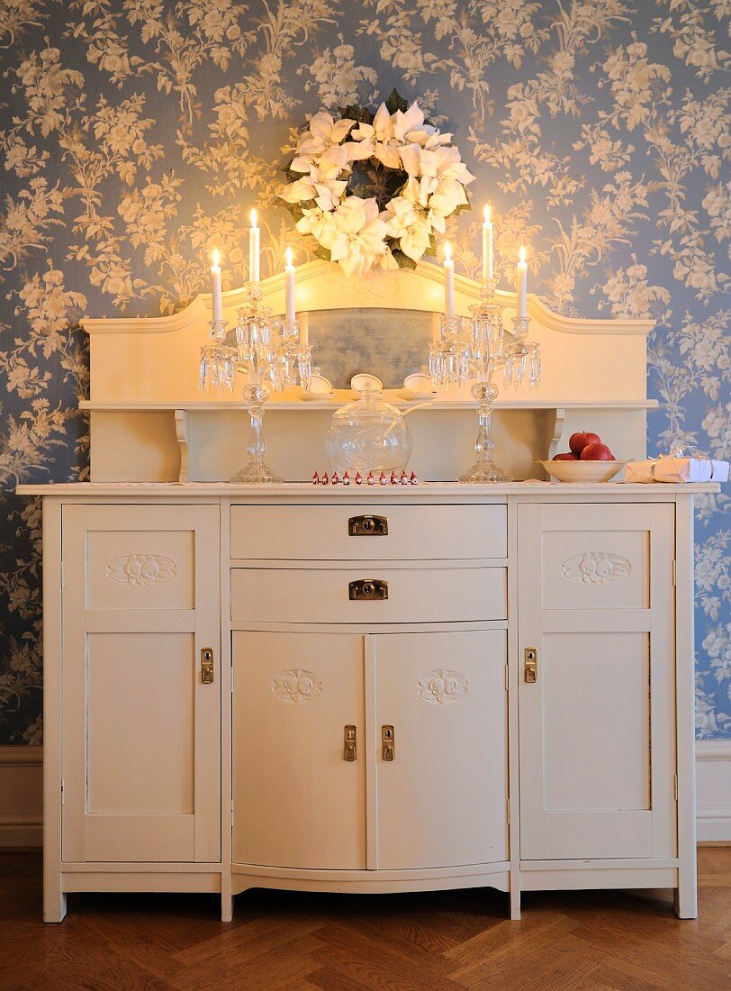 Crystal candelabra with lit candles on white-painted cabinet with top section against wallpaper with white floral pattern on blue background