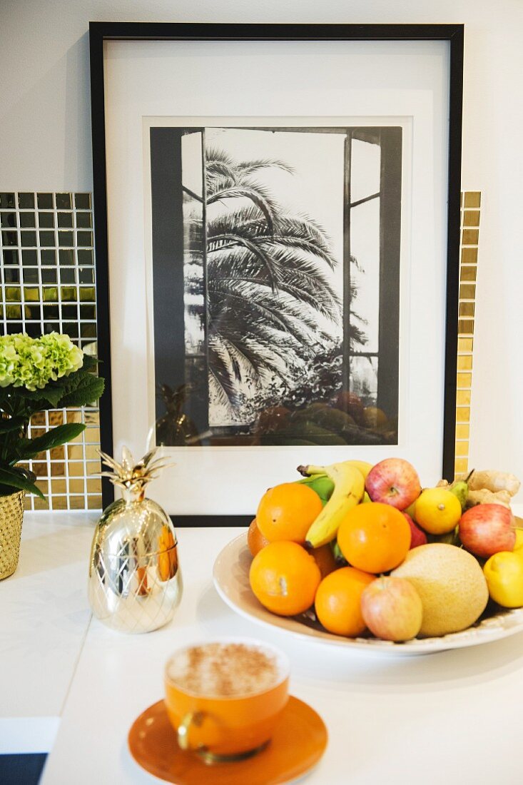 Cappuccino in orange cup and saucer in front of fruit bowl on white worksurface and framed photo leaning against mosaic splashback