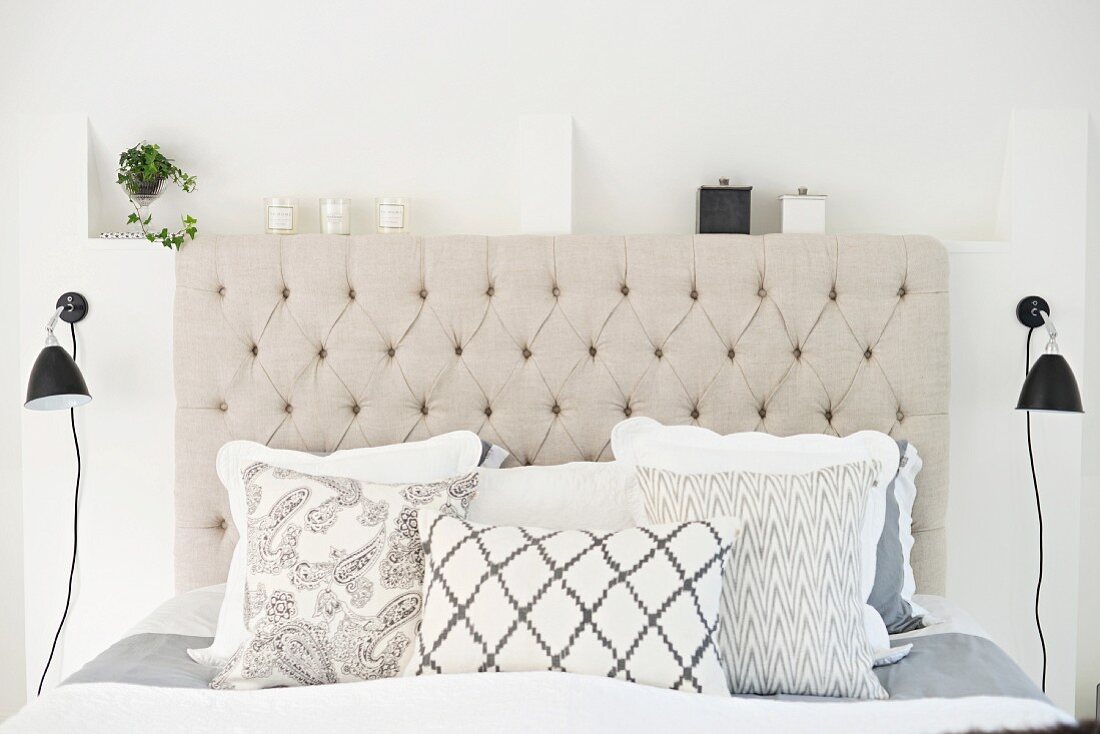 Double bed with stack of scatter cushions and button-tufted headboard in natural shades flanked by two black, retro wall lamps