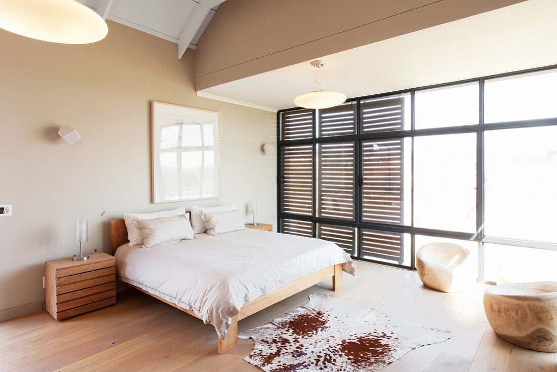 Animal-skin rug on fine wooden floor next to simple double bed in spacious bedroom with wooden, slatted sliding elements on glass wall