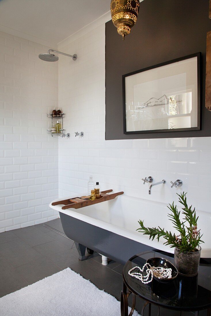 Beistelltisch mit Pflanze neben freistehender Badewanne, an Wand gerahmte Zeichnung, in Ecke offene, bodenebene Dusche in weiss gefliestem Bad