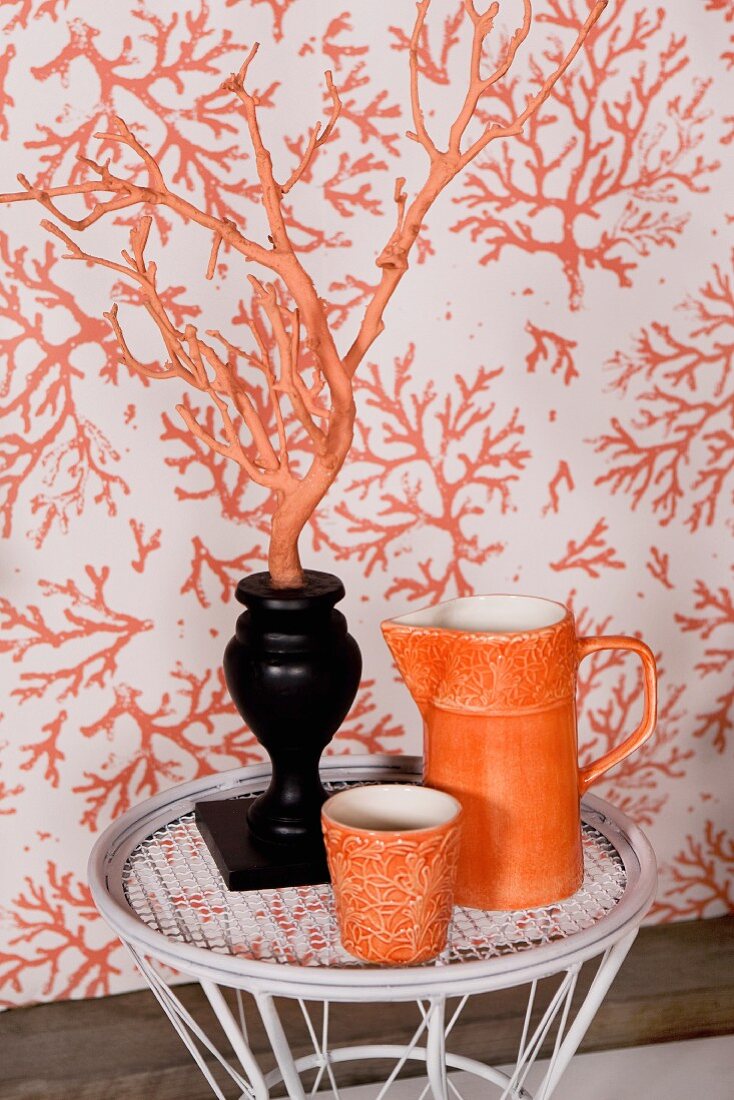 Orangefarbener Krug und Becher, neben schwarzer Vase mit orangem Dekozweig vor gemusterter Tapete