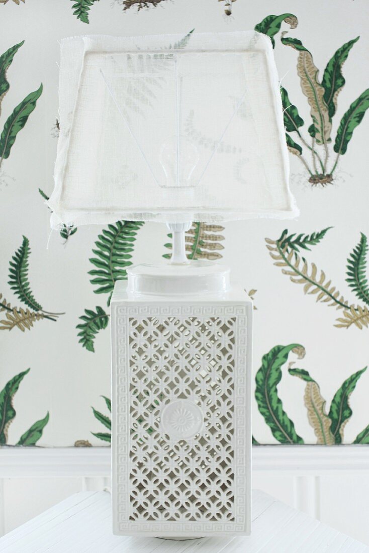 Tischleuchte mit weißem Keramikfuss und filigranem Lochmuster, transparenter Lampenschirm, im Hintergrund Tapete mit Blättermuster