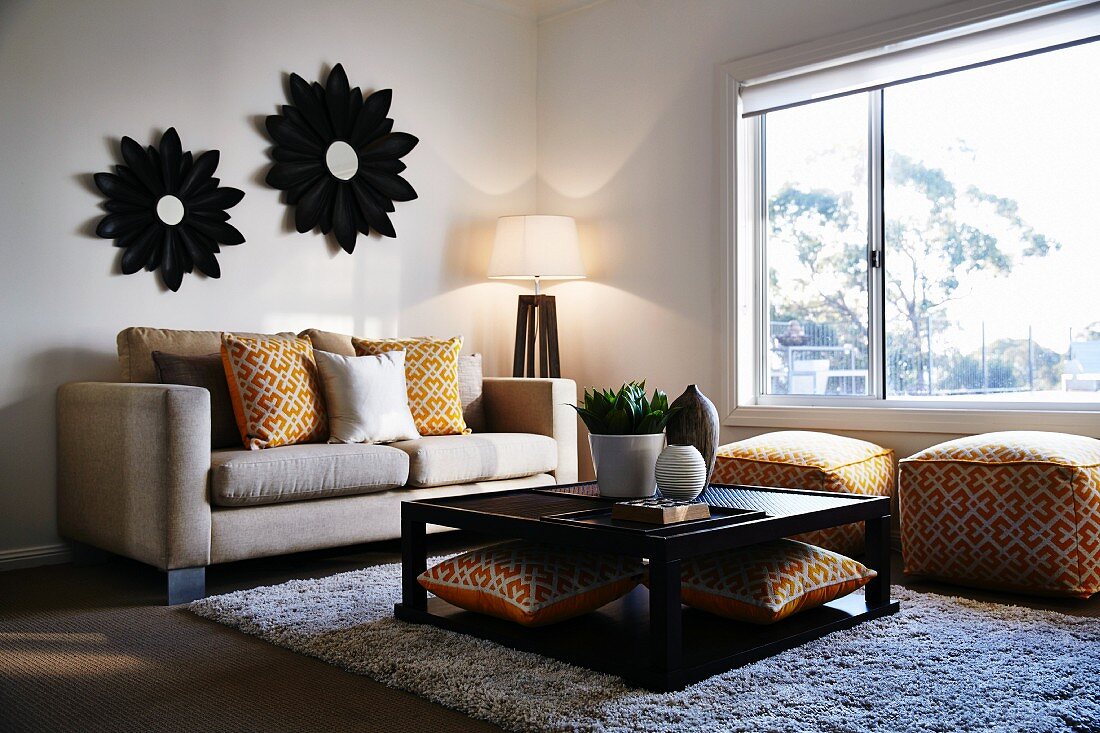 Square coffee table in front of sofa with patterned scatter cushions and matching ottomans in corner below window; mirrors with flower-shaped frames on opposite wall