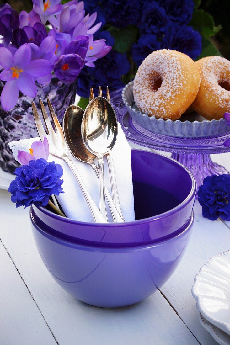 Cutlery in a purple bowl with doughnuts and purple spring flowers in the background