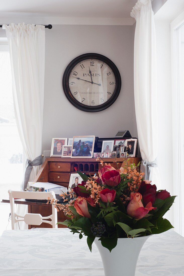 China vase of flowers on table; antique clock above writing desk in background