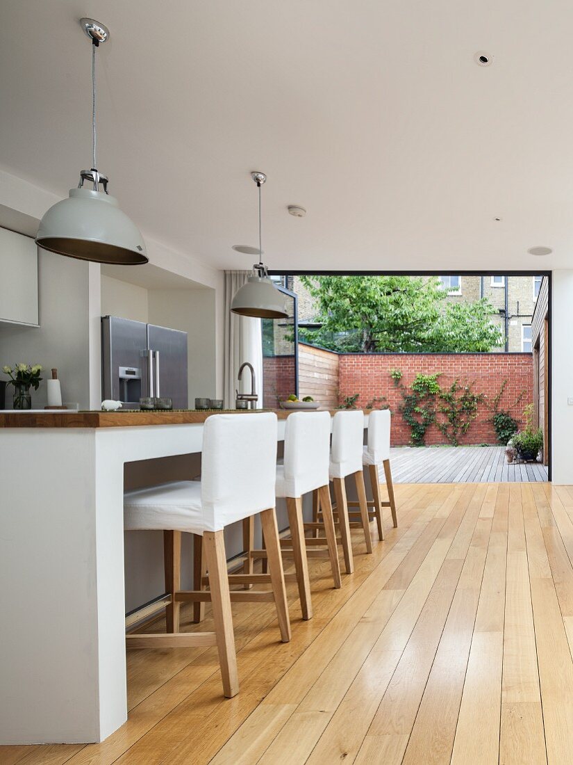 Bar stools with white loose covers at kitchen counter in open-plan designer kitchen; open folding terrace doors in background