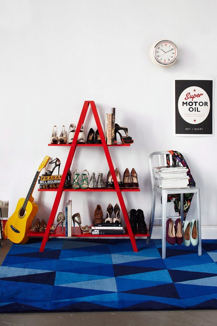 Guitar leaning against red ladder shelves of ladies' shoes and metal bar stool on rug with blue geometric pattern below poster on white wall