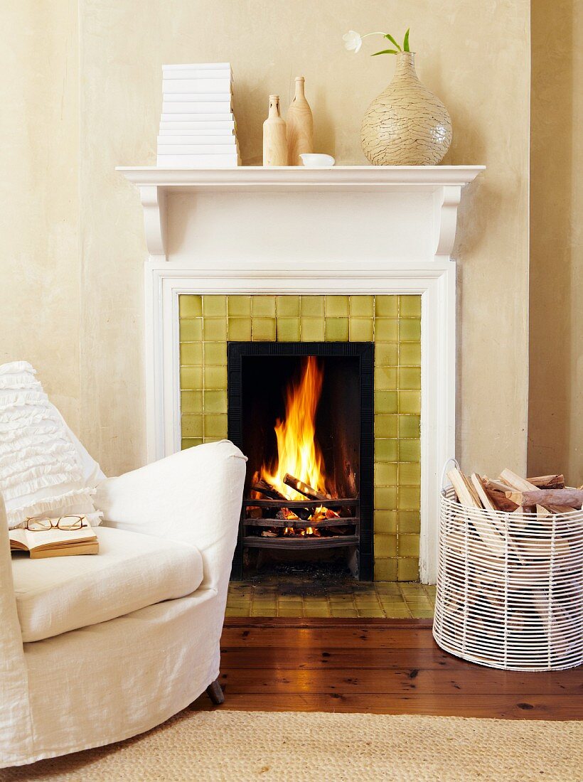 Loose-covered armchair and basket of firewood in front of blazing open fire; books wrapped in white covers and vases on mantelpiece