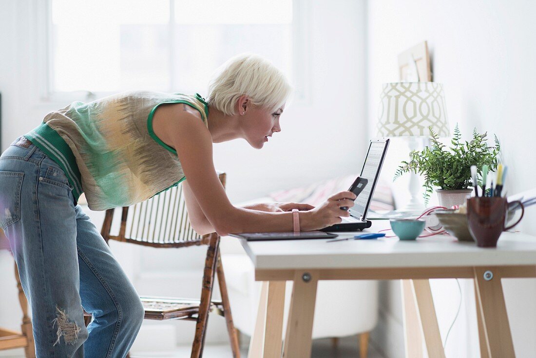 Frau mit Laptop und Handy vor dem Schreibtisch stehend