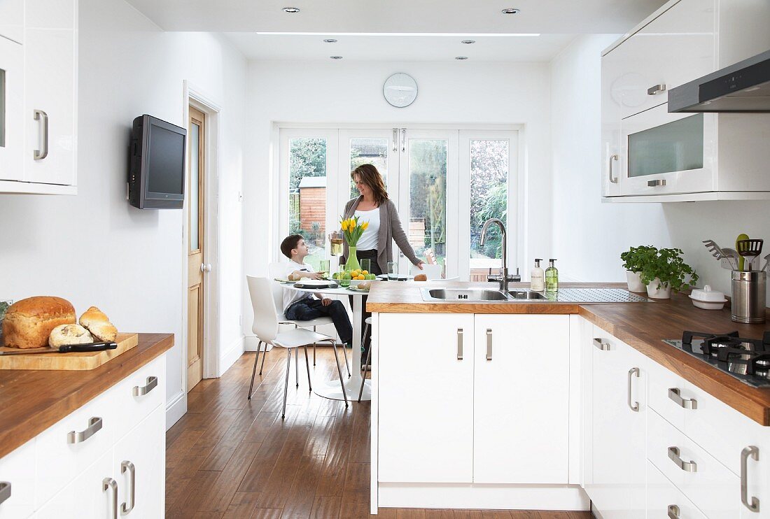 Kitchen-dining room with white fitted cupboards and wooden work surfaces; mother and son in dining area in background