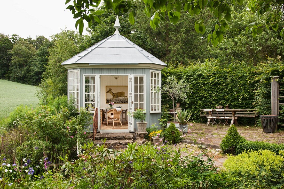 Polygoner Gartenpavillon aus hellgrau lackiertem Holz und Sprossenfenster, Blick durch offene Tür auf Sitzplatz und Bild an Wand mit Elchmotiv