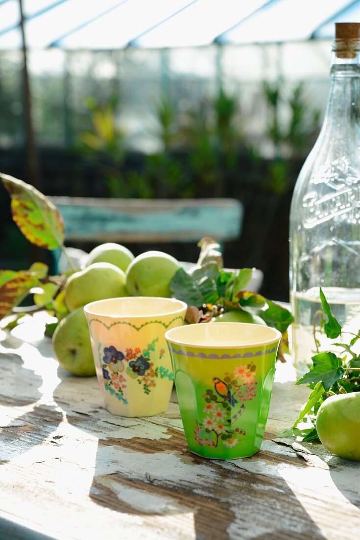 Bemalte Bechern, Apfelzweig und und Vintage Limonadenflasche auf rustikalem Tisch im Garten