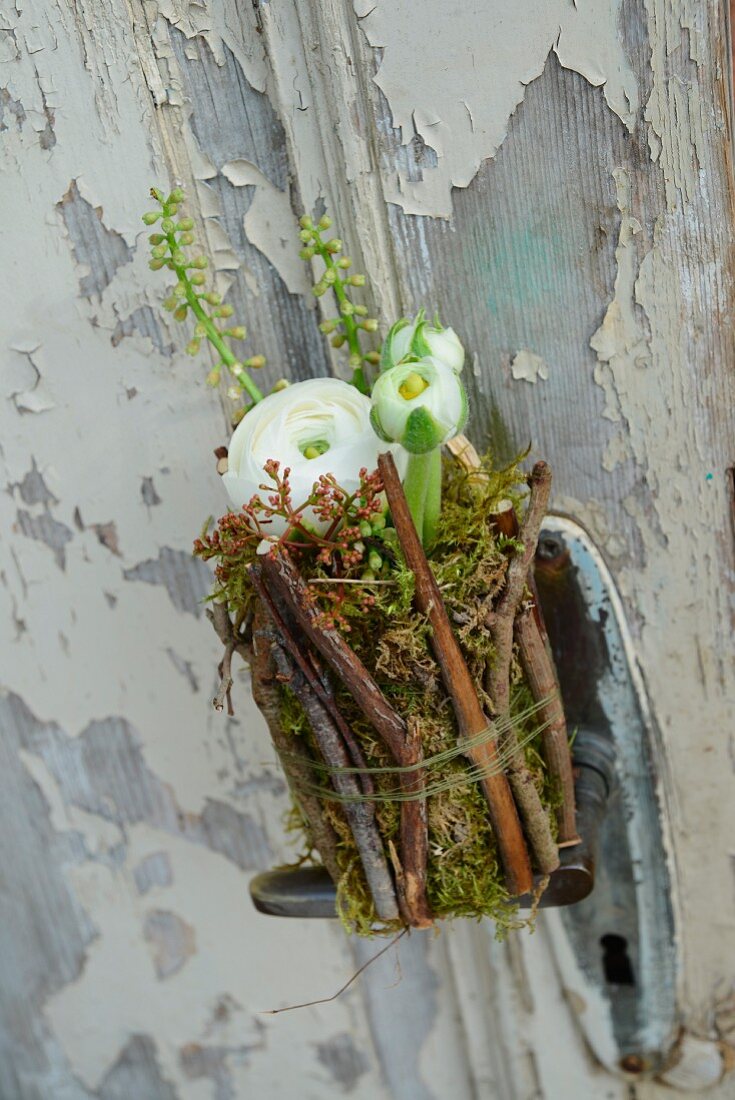 White ranunculus in jam jar covered in twigs and moss attached to door with peeling paint