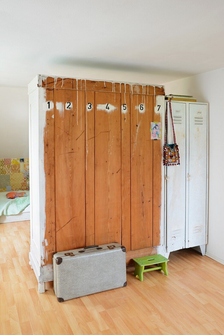 Old wardrobe and white metal lockers used as partition; numbers painted on back of wooden wardrobe with vintage suitcase and green footstool on floor