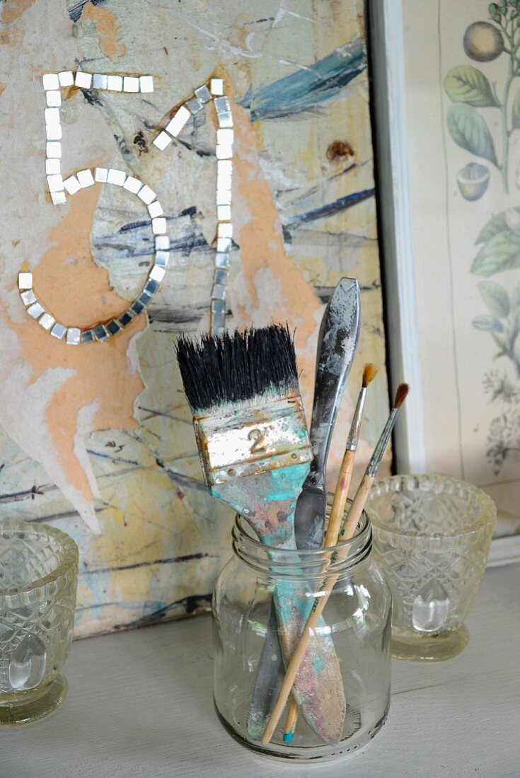 Various paintbrushes in jam jar between tealight holders in front of number made from mirrored mosaic tiles on panel