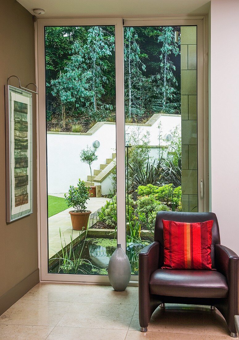 Dark brown leather armchair with red striped satin cushion in front of sliding glass door with view of garden