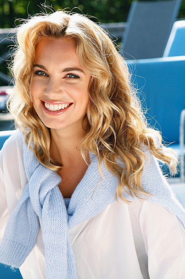 A young blonde woman outside wearing a white blouse with a jumper over her shoulders