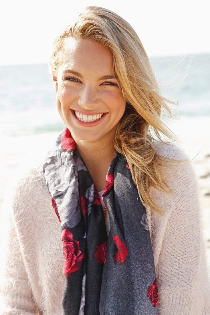 A young blonde woman by the sea wearing a light jumper and a scarf