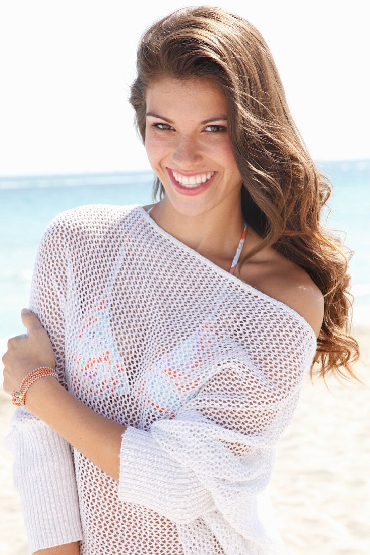 A young brunette woman on a beach wearing a white crocheted jumper