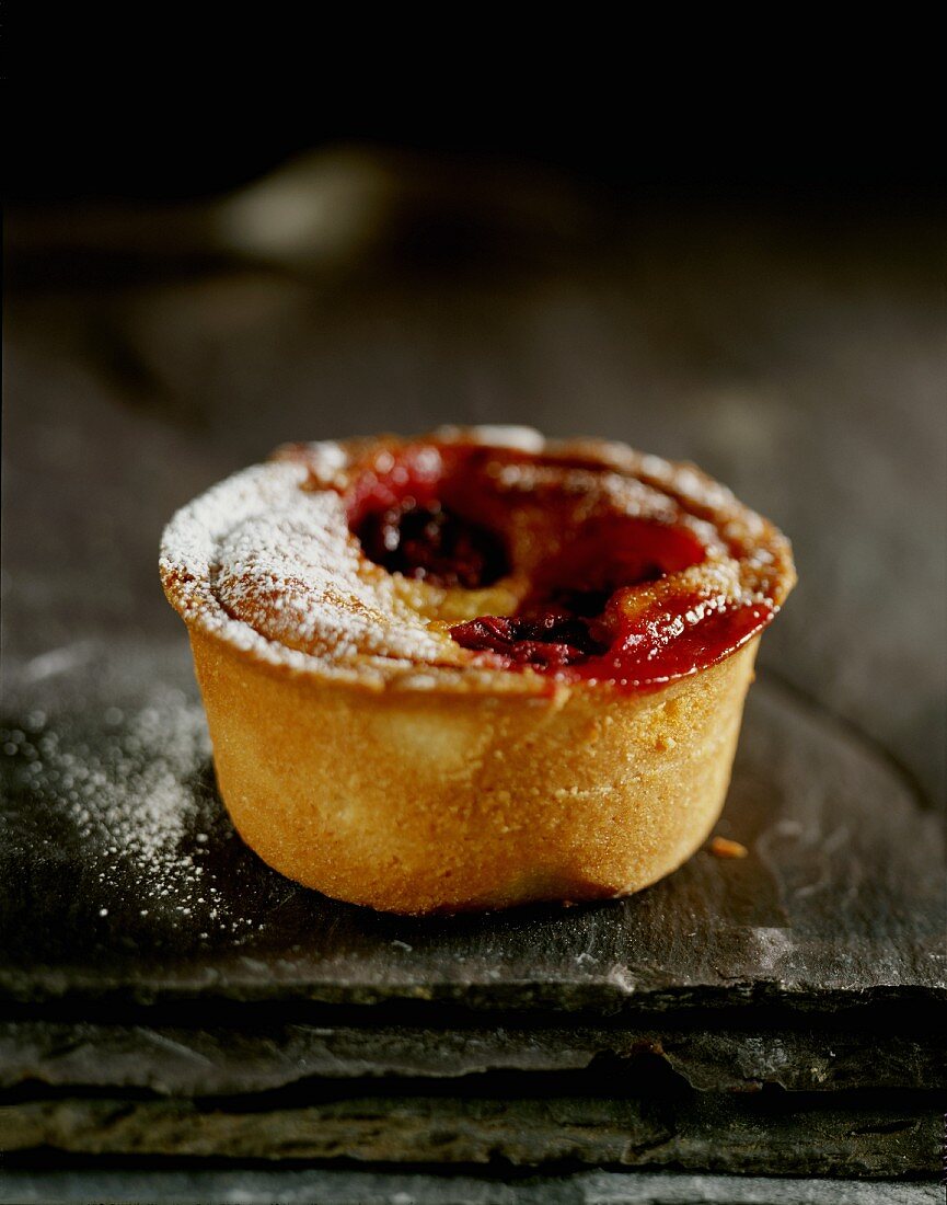 A mini cherry pie with icing sugar on a black slate