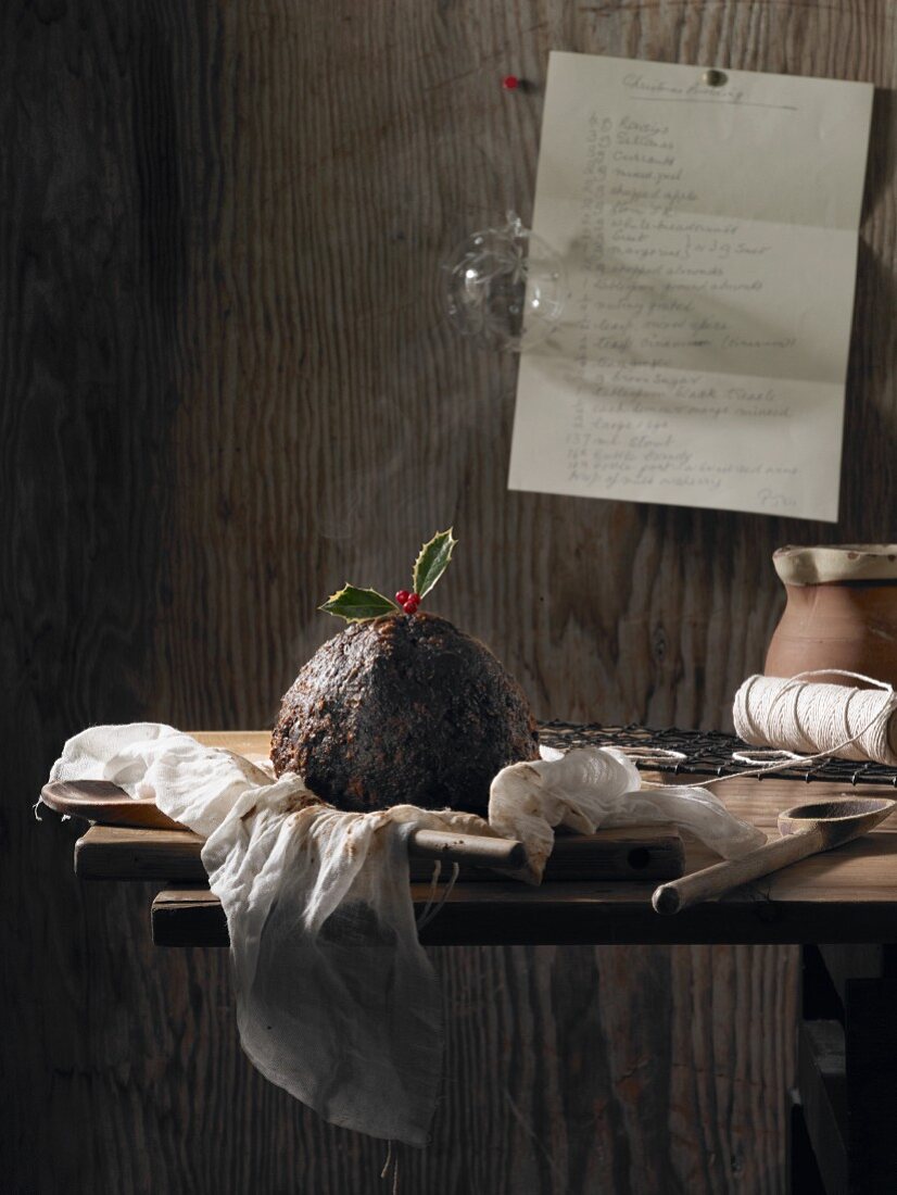 A Christmas pudding decorated with holly on a rustic wooden table with a hand-written recipe pinned to the wall behind