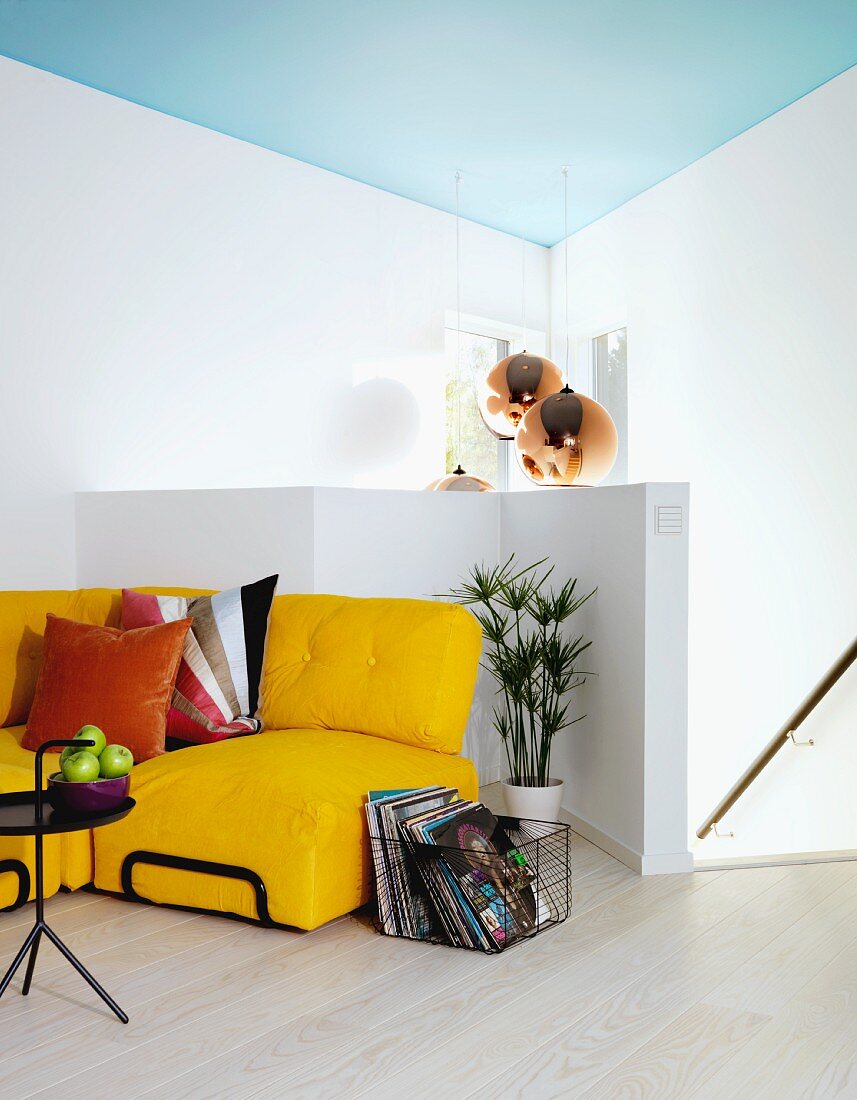 Yellow sofa with cushions next to wire basket, black metal side table, half-height white balustrade and stairs in background, pendant lamp hanging from pale blue ceiling