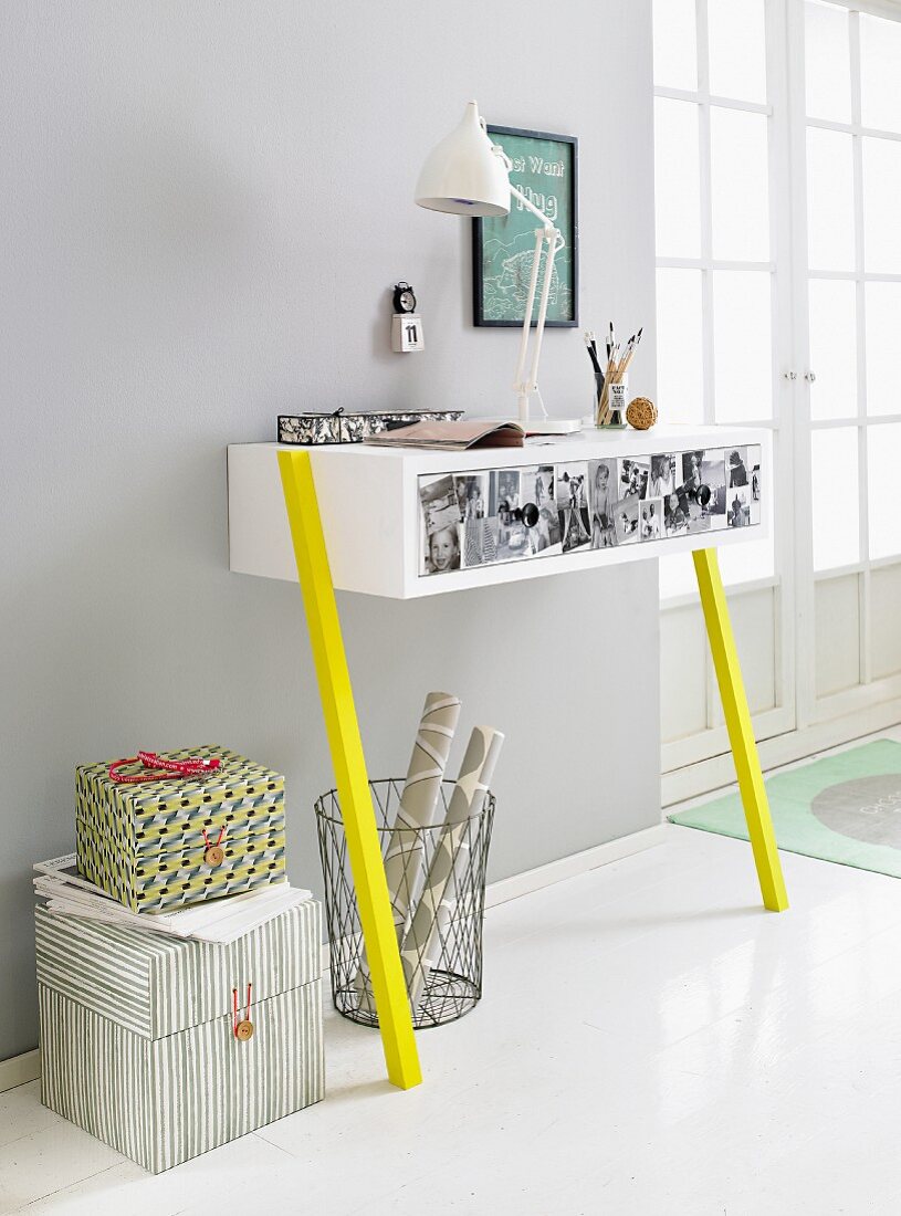 A homemade wall table against a light grey wall made from a white drawer unit decorated with photos and mounted on bright yellow legs with a waste paper basket and storage boxes underneath