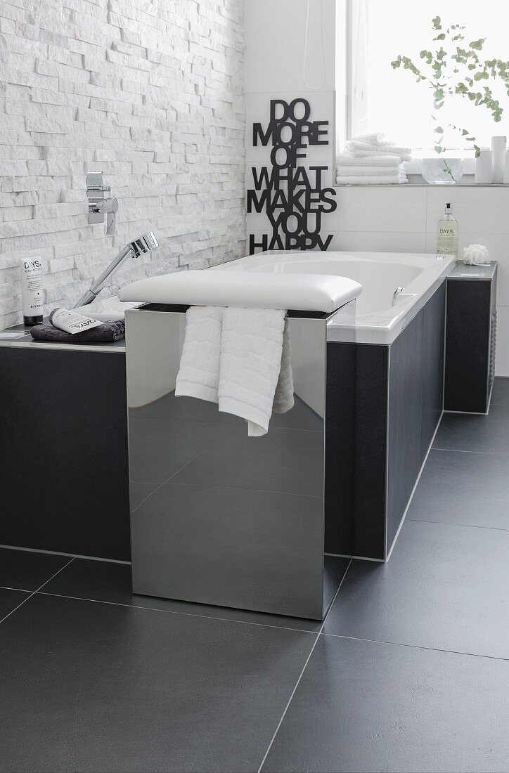 A chrome plated box with a cushion as a combined stool and laundry basket in front of a bubble jet bathtub against a natural stone wall made from decorative facing strips contrasting with the smooth, black tiles on the floor
