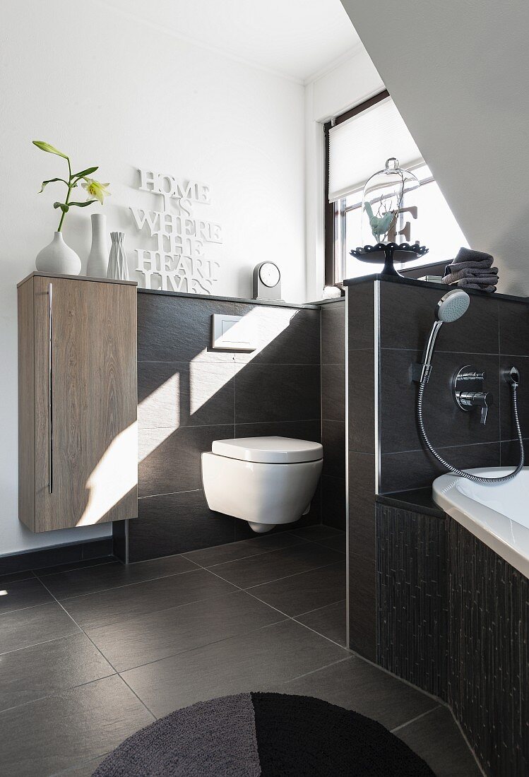 A dividing wall between a bath and a toilet, with a small wall-mounted, oak-effect cupboard next to a low tiled wall with dark brown tiles