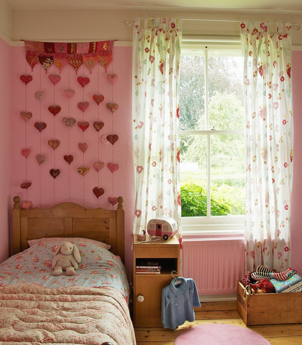 Girl's bedroom in romantic, country-house style with floral bed cover and curtains; wall hanging with heart-shaped pendants on pink wall