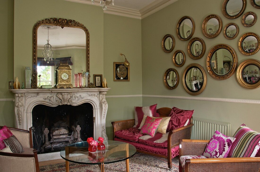 Gallery of gilt-framed, convex mirrors in interior with antique, re-upholstered seating, 60s glass table and historical fire surround