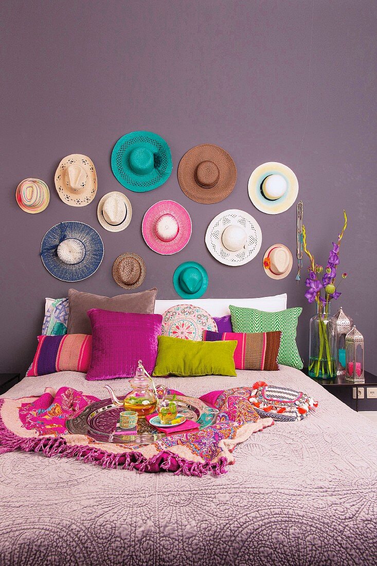 A double bed with various different coloured cushions against a purple wall hung with hats