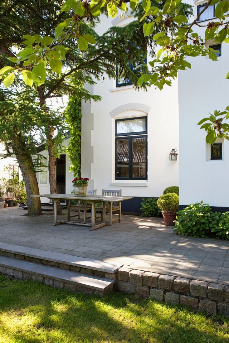 Wooden table below trees on raised terrace with steps in garden of Dutch house