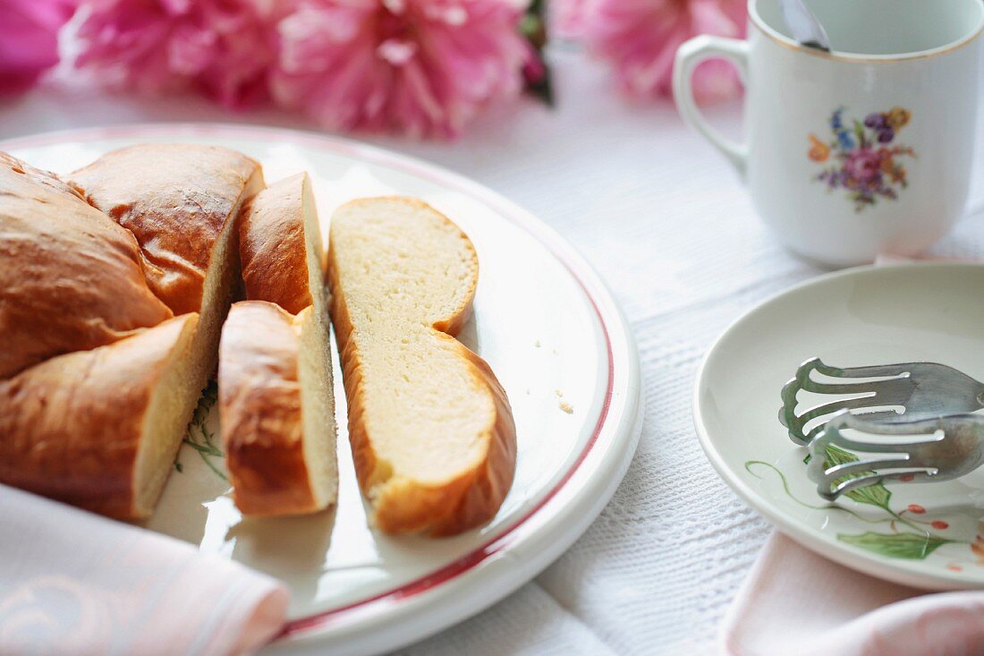 Sweet yeast bread on a rustic ceramic platter with a coffee cup, cake tones, a napkin, tablecloth and peonies