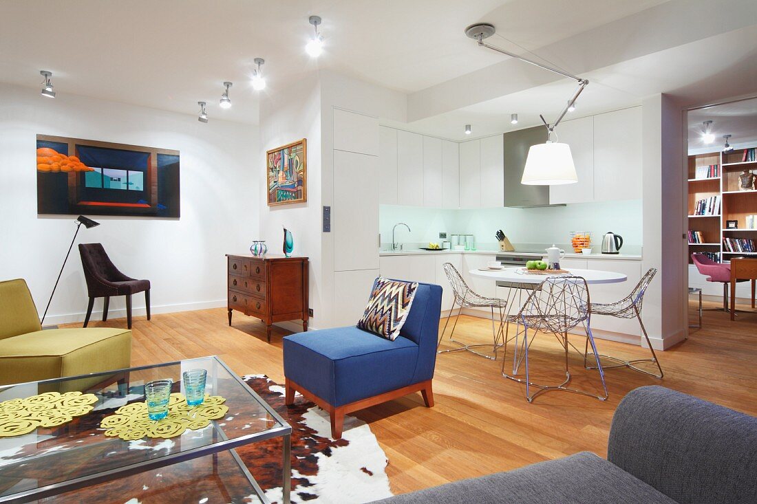 Colourful armchairs, glass coffee table, cowhide rug and wooden floor; dining area with wire chairs and open-plan kitchen in background