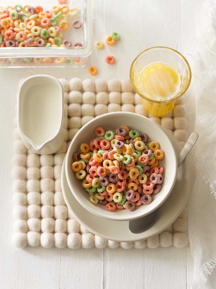 Bunte fruchtige Cerealienkringel im Schälchen, Milchkännchen und ein Glas Orangensaft