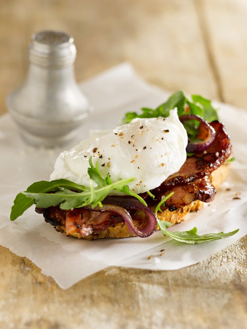 A slice of bread topped with a poached egg, bacon and rocket