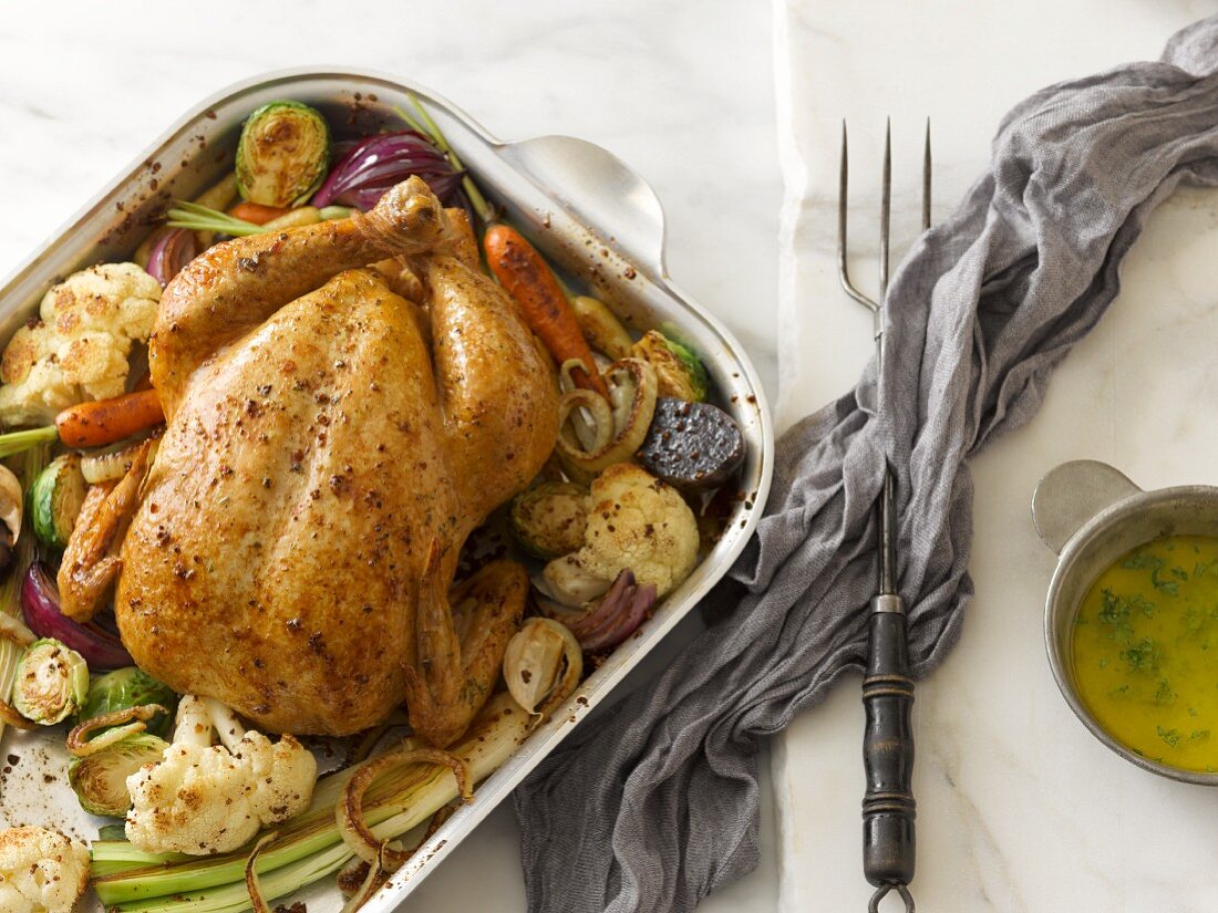 A whole roast chicken in a roasting tin surrounded by cauliflower, carrots, Brussels sprouts and onions with herb butter on the side