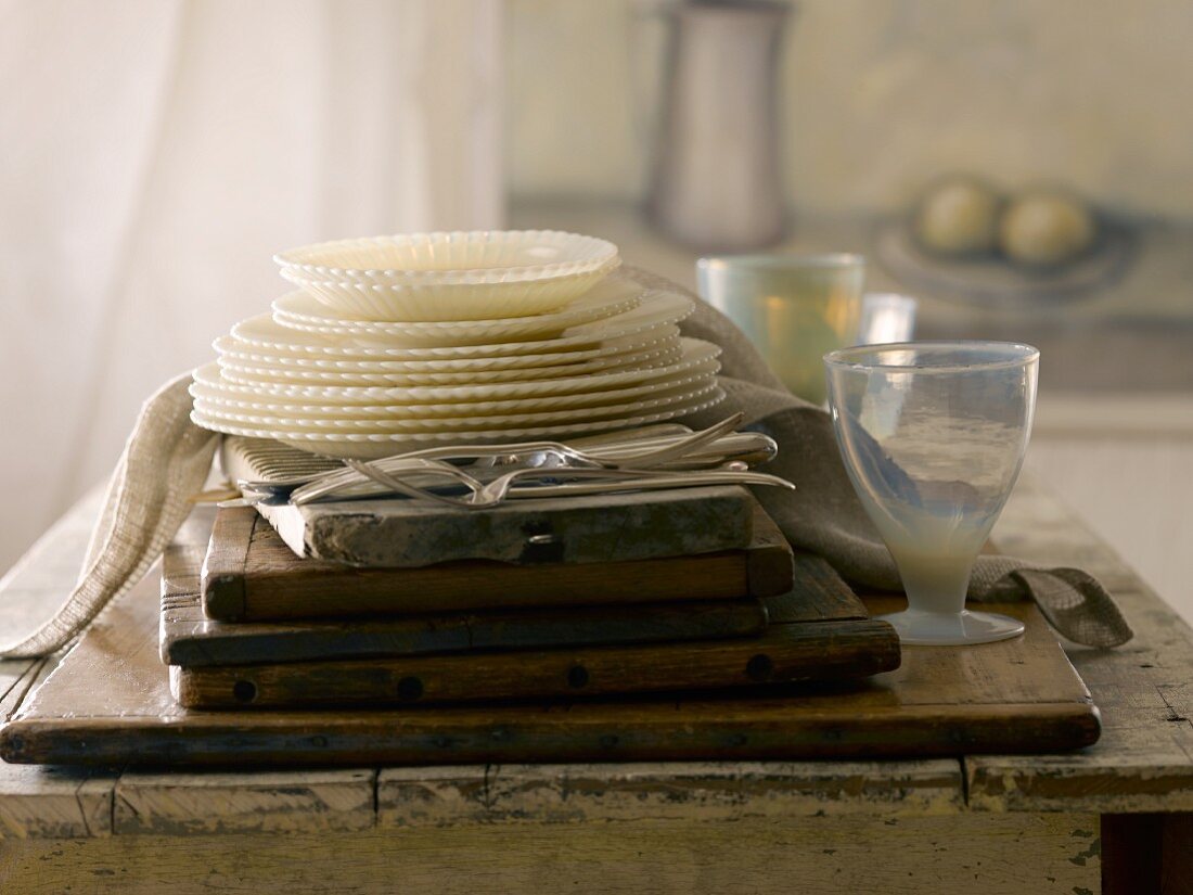 A stack of vintage plates, glassware and cutlery in front of a painting and a curtain