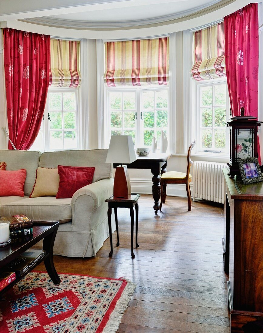 Wohnzimmer mit Erker, an Sprossenfenster gestreifte Rollos und pinkfarbene Vorhänge, im Vordergrund teilweise sichtbare Couch und antiker Beistelltisch