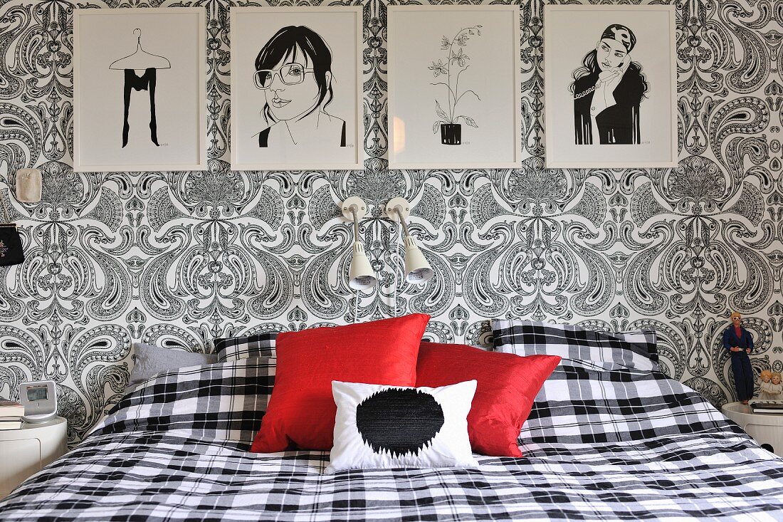 Schlafzimmer mit schwarz-weissen Zeichnungen, floralem Paisleymuster und karierter Bettwäsche, Farbakzent mit roten Kissen
