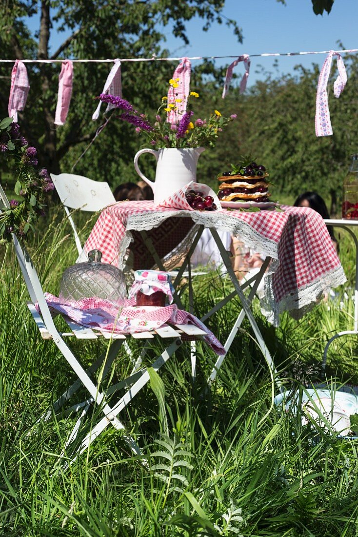 Gartentisch mit karierter Decke, darauf frische Kirschen und Krug mit Wiesenblumen in sonnenbeschienenem Garten