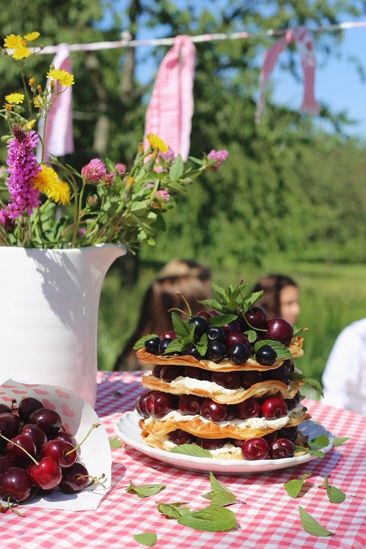 A stack of waffle with cream and fresh cherries on a plate next to a jug of wild flowers on a checked tablecloth