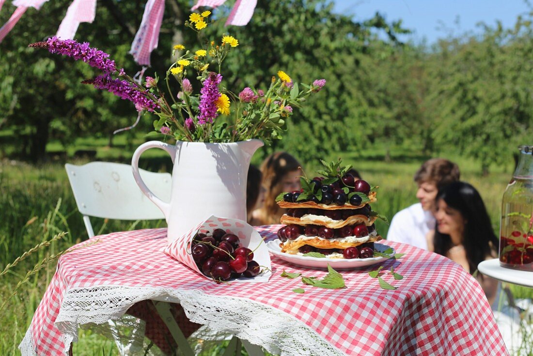 Frische Kirschen, Waffelturm und Wiesenblumen in weißem Krug auf Gartentisch mit karierter Tischdecke, im Hintergrund Menschen