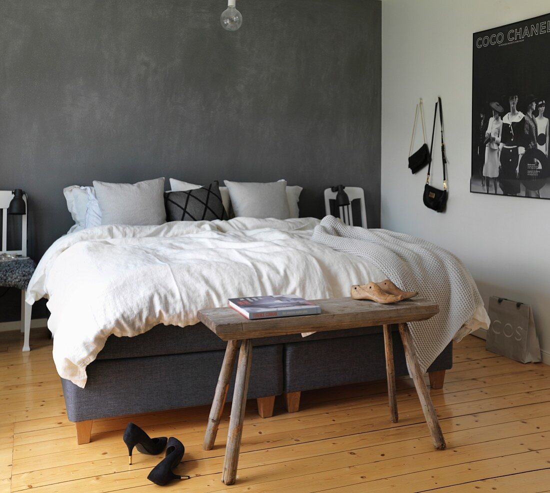 Doppelbett vor grauer Wand, naturbelassener Dielenboden und rustikale Kleiderbank im Schlafzimmer
