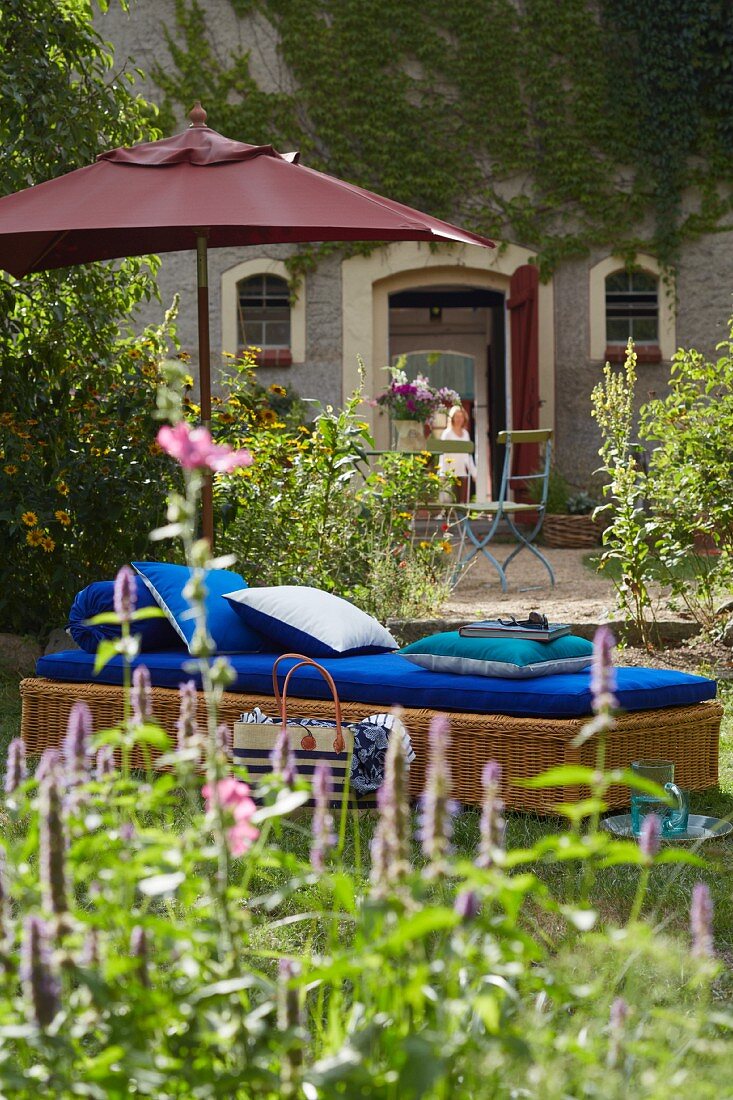 A parasol and a wicker lounger with blue cushions in a garden with a vine-covered facade of a French country house in the background